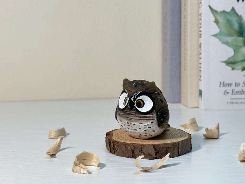 Puede incluir: Una peque&ntilde;a figura de b&uacute;ho tallada a mano en madera con detalles pintados en marr&oacute;n y blanco, que presenta grandes ojos con borde blanco, iris amarillos y pupilas negras, posada sobre una rodaja de madera circular con corteza natural.