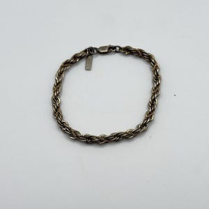 May include: Silver-toned rope chain bracelet. The bracelet features a twisted rope design and a small rectangular tag near the clasp. The bracelet is displayed on a white background.