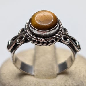 May include: A silver ring featuring a round, polished tiger's eye gemstone set in a bezel. The setting is surrounded by a decorative twisted rope border, and the band displays intricate scrollwork details on both sides of the central stone.
