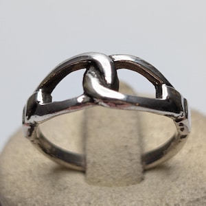 May include: A polished silver-toned ring featuring a central design of two interlocking, curved metal links that form a continuous, open-work pattern. The band is smooth and uniform, showcasing a reflective metallic finish throughout the entire piece.