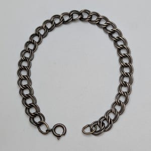 May include: A dark silver-toned metal bracelet featuring a double-link chain design. The bracelet is constructed with interconnected circular links and is secured with a round spring-ring clasp mechanism at one end of the chain.