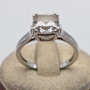 Op de afbeelding: Een zilverkleurige ring met een grote, heldere edelsteen in kussenslijpvorm, vastgehouden door vier pootjes. De ringband is glad en gepolijst, met een eenvoudig en strak ontwerp dat de centrale steen en het gefacetteerde oppervlak benadrukt.