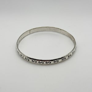 Puede incluir: Un brazalete de plata con un patrón repetitivo de espirales estilizadas. El brazalete circular es de metal. El diseño está grabado en la superficie. Una joya clásica.