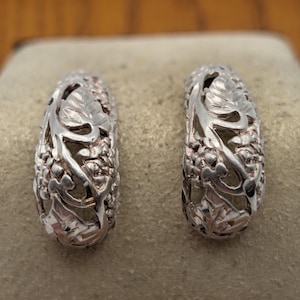 Peut inclure: Une paire de boucles d'oreilles créoles argentées présentant un design ajouré complexe composé de feuilles en relief et de petites grappes de baies. Le métal possède une finition polie et réfléchissante avec des découpes détaillées.