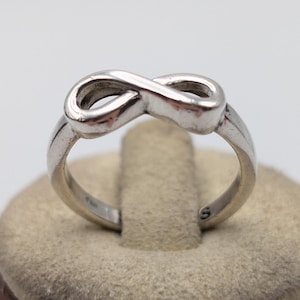 May include: A polished silver-toned ring featuring a prominent infinity symbol design centered on the band. The metal surface is smooth and reflective, showcasing a continuous loop shape that forms the focal point of this jewelry piece.