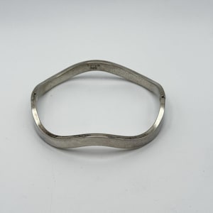 May include: A silver-toned, wavy bangle bracelet. The bracelet has a simple, elegant design with a subtle, undulating shape. The metal has a polished finish, reflecting light. The bracelet is a piece of jewelry.
