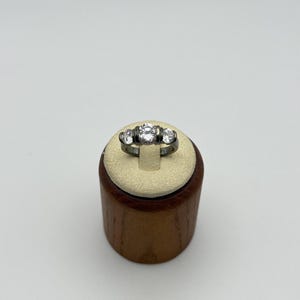 May include: A silver ring with three clear, round gemstones. The center stone is larger than the two flanking stones. The ring is displayed on a small, wooden ring holder with a cream-colored top.