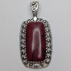 May include: A rectangular pendant featuring a smooth, deep maroon stone with dark veining, set within a polished silver frame. The metal border displays intricate scrollwork and a twisted rope detail surrounding the central stone, topped with a decorative silver bail.