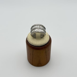 May include: A silver ring with two rows of rectangular-cut stones and a smaller row of round stones. The ring is displayed on a small, round wooden stand with a beige top.
