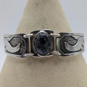 May include: A silver cuff bracelet featuring a central oval dark gray stone set in a rectangular frame. The bracelet has two side panels connected by silver loops, each displaying a smooth, raised bird silhouette against a textured background.