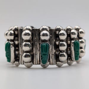 Könnte beinhalten: Ein breites Armband aus Silber mit drei vertikalen Reihen aus polierten Silberkugeln und drei eingesetzten, grün geschnitzten Gesichtern aus Stein. Die Metalloberfläche hat ein angelaufenes Finish mit vertikalen Linien zwischen den Reihen.