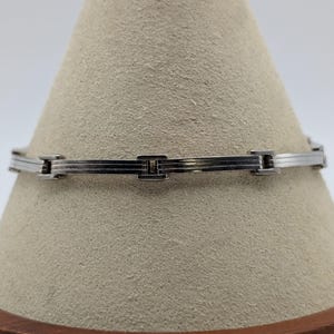 May include: A silver-toned metal link bracelet featuring a series of rectangular segments connected by small, polished hinges. The bracelet has a smooth, reflective finish with subtle horizontal lines etched into each individual metal link.