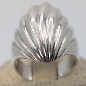 May include: A polished silver ring featuring a prominent, raised shell-like design with vertical fluted ridges. The band is wide and tapers toward the back, showcasing a smooth, reflective metallic surface with a curved, scalloped top edge.