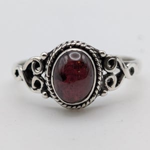 May include: A silver ring featuring a central oval-shaped dark red gemstone. The stone is set within a twisted rope-style metal border, flanked by decorative scrollwork patterns on both sides of the band, creating a detailed metalwork design.