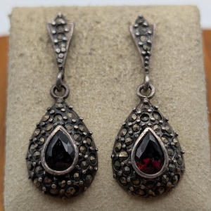 May include: A pair of drop earrings featuring a teardrop-shaped dark red gemstone center surrounded by small, metallic, textured accents. The earrings have a pointed, triangular top section connected by a small ring to the main body.