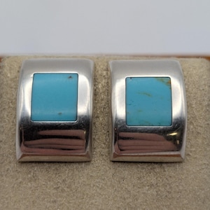 Puede incluir: Un par de pendientes de plata pulida, cada uno con un marco de metal rectangular curvado con una incrustación cuadrada central de piedra turquesa azul brillante. La superficie metálica lisa refleja la luz, resaltando el contraste entre ambos.