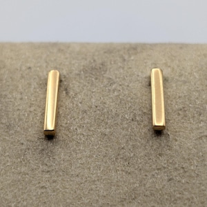 May include: A pair of gold-toned stud earrings, each featuring a simple, smooth, vertical rectangular bar design. The earrings have a polished metallic finish and are presented in a clean, straightforward orientation, highlighting their minimalist shape.