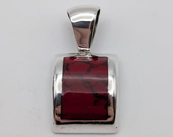 May include: A rectangular pendant featuring a polished silver frame and a central inlay of deep red stone with dark, web-like veining. The piece includes a large, smooth silver bail at the top for attachment to a chain or cord, creating a sleek look.