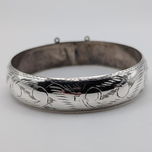 May include: A silver-toned hinged bangle bracelet featuring a polished finish and intricate engraved scrollwork patterns across the outer surface. The circular band includes a secure clasp mechanism and two small loops at the top edge.