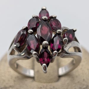 May include: A silver ring featuring a cluster of seven marquise-cut deep red garnet gemstones arranged in a vertical, pointed formation. The stones are held in place by small metal prongs, set upon a polished silver band with openwork sides.