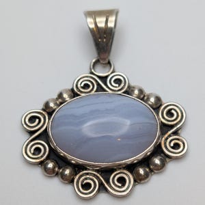 May include: An oval-shaped pendant featuring a smooth, light blue banded stone set in a sterling silver frame. The silver border is decorated with intricate metal swirls and small spherical beads, topped with a triangular silver bail for hanging.