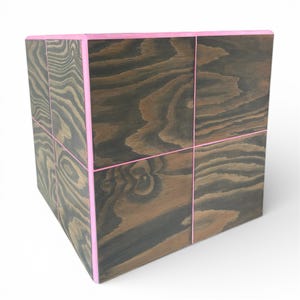 Könnte beinhalten: Ein Holzwürfel mit dunkelbraunem Finish und rosa Akzenten. Der Würfel ist durch rosa Linien in Abschnitte unterteilt, wodurch ein gitterartiges Muster entsteht. Die Holzmaserung ist sichtbar und verleiht Textur.