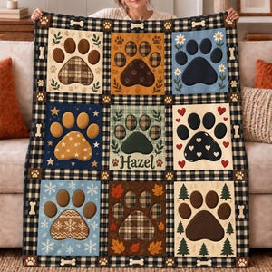 May include: A rectangular throw blanket featuring a black and white buffalo plaid border with bone and paw print accents. The center contains nine square panels, each displaying a large paw print with unique patterns, including plaid, hearts, snowflakes, leaves, and trees. One panel features the name Hazel.