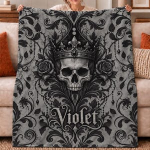 Peut inclure: Une couverture rectangulaire en polaire présentant un motif damassé gris et noir avec un crâne central portant une couronne, flanqué de deux roses. Le prénom Violet est imprimé dans une police de style gothique au bas de la couverture.