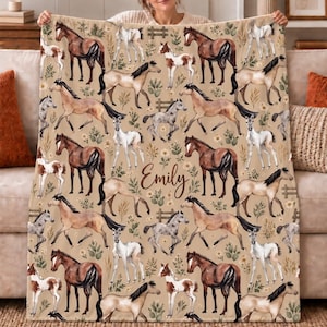 Könnte beinhalten: Eine beige Wohndecke mit einem sich wiederholenden Muster aus aquarellartigen Pferden und Fohlen in verschiedenen Braun-, Weiß- und Grautönen, durchsetzt mit kleinen grünen Pflanzen und Blumen. Der Name Emily ist mittig in brauner Schreibschrift gedruckt.