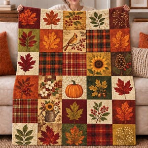 May include: A rectangular quilt featuring a patchwork design of twenty-five squares. The squares display autumn-themed imagery including maple leaves, a cardinal on a branch, plaid patterns, a sunflower, a pinecone, a pumpkin, and a watering can.