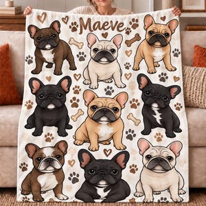 Puede incluir: Una manta de forro polar blanca con un patrón repetitivo de bulldogs franceses de dibujos animados en colores marrón, negro y crema. El diseño incluye huellas, huesos y corazones marrones, con el nombre Maeve impreso en texto marrón.