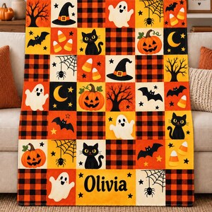 May include: A rectangular throw blanket featuring a grid pattern of Halloween-themed squares in orange, black, and white. The design includes ghosts, black cats, pumpkins, bats, spiderwebs, and plaid squares. The name Olivia is printed in black.