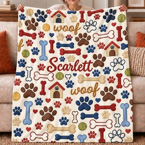 May include: A cream-colored throw blanket featuring a repeating pattern of dog-themed icons including brown, blue, and red paw prints, bones, tennis balls, and dog houses. The blanket includes the name Scarlett and the word woof in script.