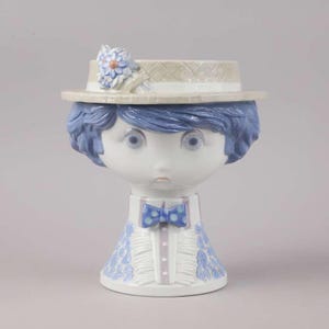 May include: A ceramic figurine depicting a face with blue hair and eyes, wearing a hat with a flower and a blue polka dot bow tie. The figurine has a white base and a blue and white patterned dress.