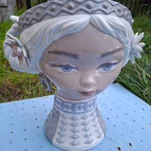 May include: A ceramic planter shaped like a woman's head, featuring a gray and white color scheme. The planter has blue eyes, a decorative crown, and floral accents. The neck area is adorned with a patterned design.