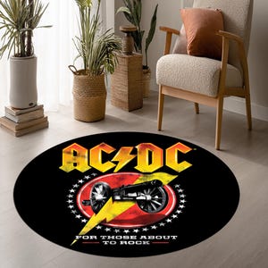 以下が含まれることがあります： 黄色とオレンジ色のAC/DCロゴが入った黒い円形ラグ。黄色の稲妻がキャノンのグラフィックの後ろにあります。「FOR THOSE ABOUT TO ROCK」というテキストが下にあります。ラグは明るい色の床にあります。
