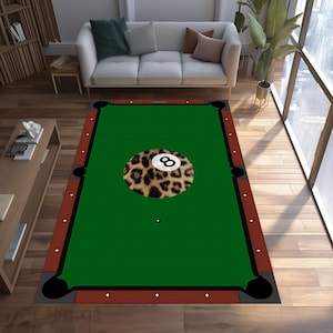 May include: A rectangular area rug designed to resemble a pool table. The rug features a green playing surface, a brown border, and black corner pockets. A large, round, leopard-print design with the number 8 is in the center.