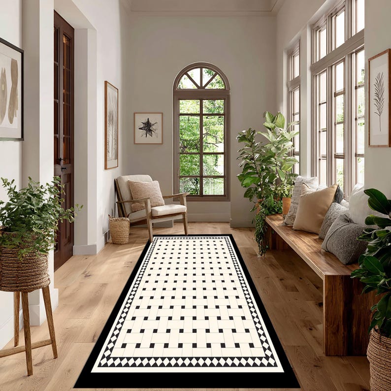 May include: A rectangular rug with a black border and a cream-colored center featuring a grid pattern of black squares. The rug is placed on a light wood floor in a hallway, with a wooden bench and plants visible.