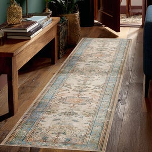 Puede incluir: Una alfombra rectangular de aspecto envejecido, con un centro crema y un borde en azul claro y turquesa. La alfombra presenta un patrón descolorido de estilo vintage con toques de naranja y marrón, ideal para un pasillo.