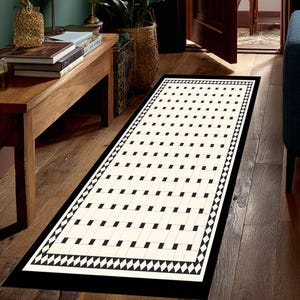 May include: A rectangular rug with a black border and a repeating pattern of black squares on an off-white background. The border features a diamond pattern. The rug is placed on a hardwood floor.