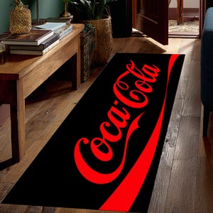 Könnte beinhalten: Schwarzer rechteckiger Teppich mit dem Coca-Cola-Logo in Rot. Das Logo ist in der klassischen Schriftart mit einem roten, geschwungenen Streifen am unteren Rand. Der Teppich liegt auf einem Holzboden.