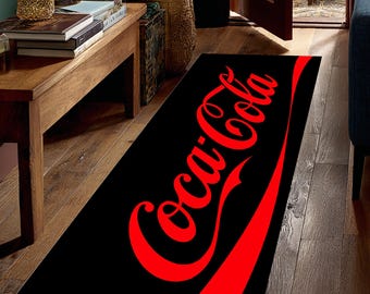 May include: Black rectangular rug featuring the Coca-Cola logo in red. The logo is in the classic script font with a red curved stripe at the bottom. The rug is placed on a wooden floor.
