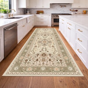 May include: A rectangular area rug with an intricate floral and geometric pattern. The rug features a cream-colored central field with accents of brown, beige, and sage green. The border is a sage green with floral motifs. The rug is placed in a kitchen.