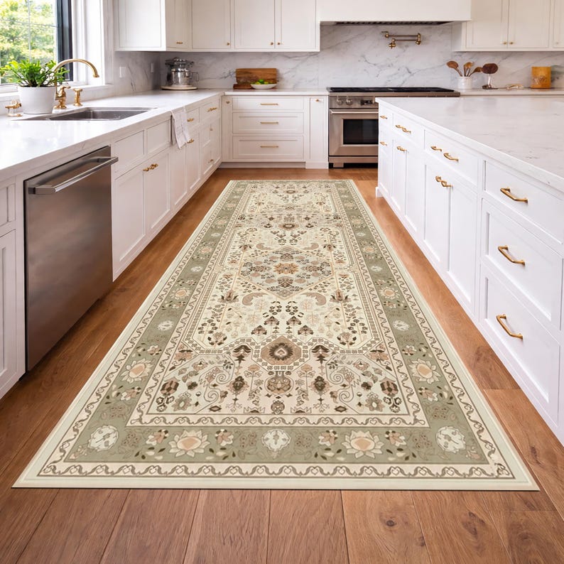 May include: A rectangular area rug with an intricate floral and geometric pattern. The rug features a cream-colored central field with accents of brown, beige, and sage green. The border is a sage green with floral motifs. The rug is placed in a kitchen.
