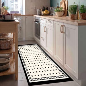 Op de afbeelding: Een rechthoekig keukenkleed met een zwarte rand en een herhalend patroon van zwarte vierkanten op een crèmekleurige achtergrond. Het kleed ligt voor witte kasten en een fornuis en voegt een decoratief element toe aan de keuken.