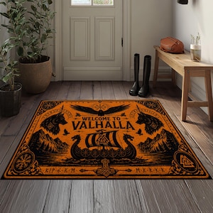 Könnte beinhalten: Eine rechteckige orange-schwarze Fußmatte mit einem Wikinger-Motiv. Das Design zeigt ein zentrales Langschiff, zwei Wolfsköpfe, einen Raben, Berge und den Text "WELCOME TO VALHALLA" zusammen mit nordisch inspirierten Symbolen und Runen.