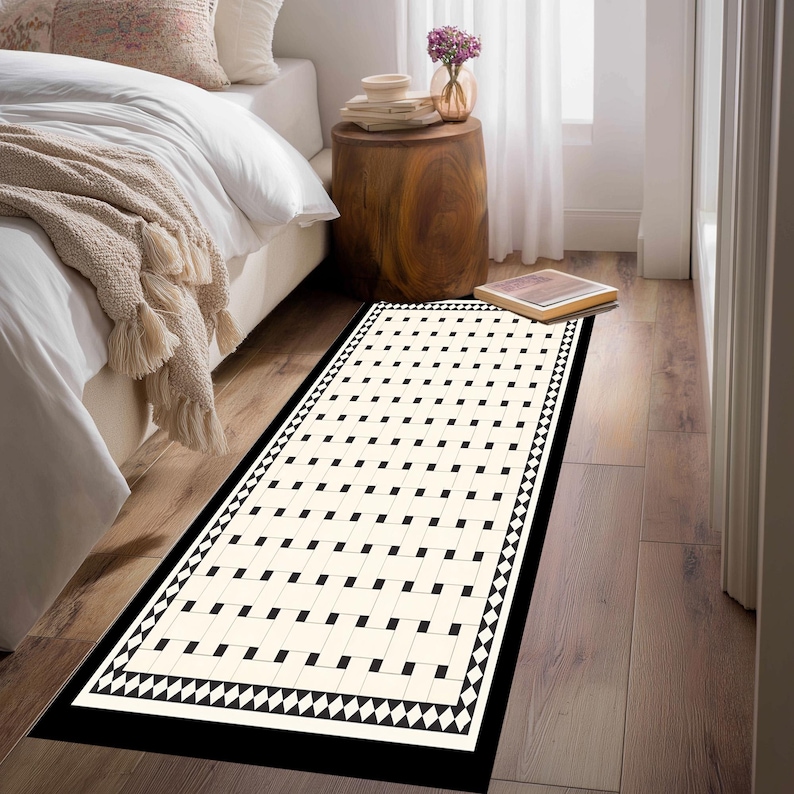 May include: A rectangular rug with a cream-colored background and a repeating pattern of black squares. The rug has a black border with a geometric diamond design. The rug is placed on a wooden floor near a bed.