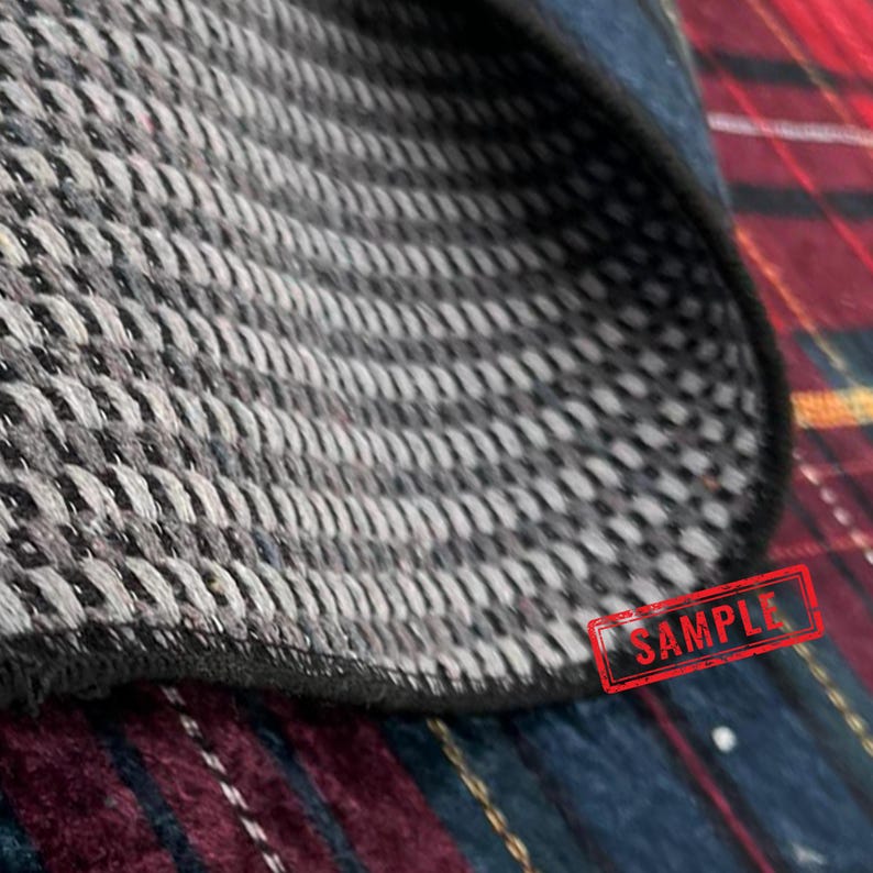 May include: Close-up of a gray and black woven fabric with a textured pattern. The fabric has a dark border and is partially covering a red, blue, and black plaid pattern. A red "SAMPLE" stamp is visible.