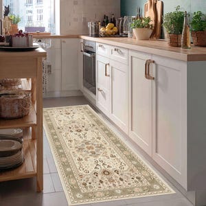 May include: A rectangular beige kitchen rug with an intricate floral pattern and a sage green border. The rug is placed in front of white cabinets and a stove, adding a decorative element to the kitchen.
