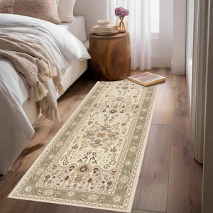 May include: A rectangular beige rug with an intricate floral pattern and a sage green border. The rug is placed on a hardwood floor, adding a touch of elegance to the room. The rug's dimensions are approximately 3 feet wide and 8 feet long.